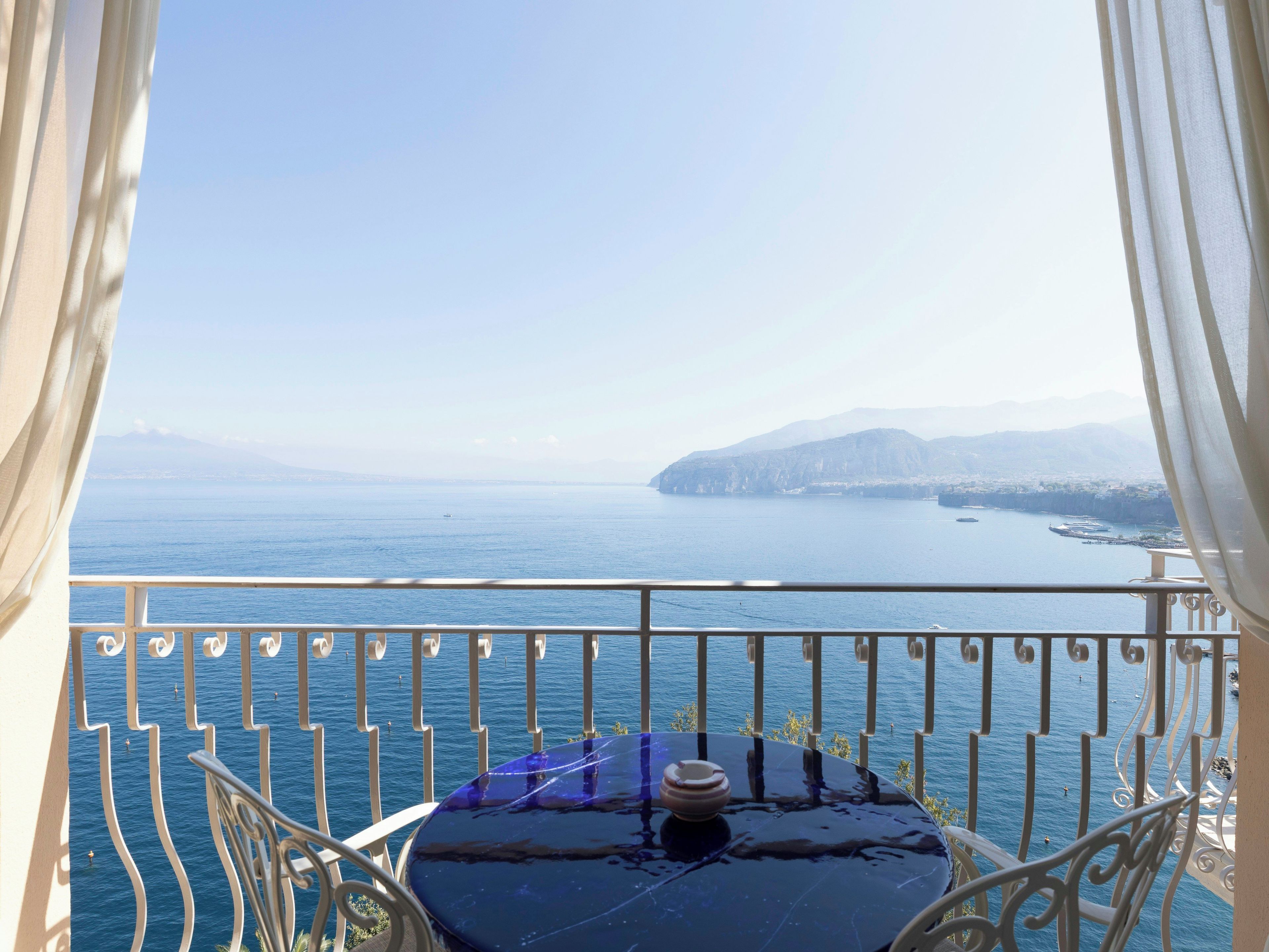 A balcony with a table and a view of the sea and distant mountains.