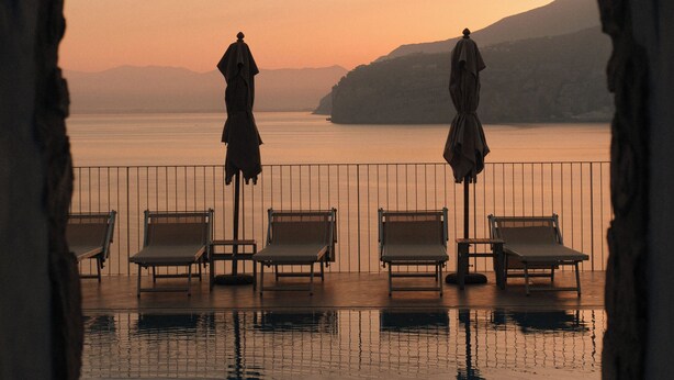 Une piscine à débordement, dotée de chaises longues et de parasols, que l’on aperçoit à travers une porte en arc, et qui offre une vue sur une étendue d’eau et sur la terre ferme au loin.
