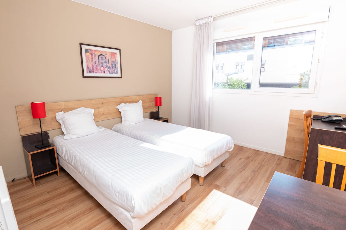 A hotel room with two single beds, a wooden headboard, a nightstand with a red lamp, a framed picture on the wall, and a window with a view of the outside.