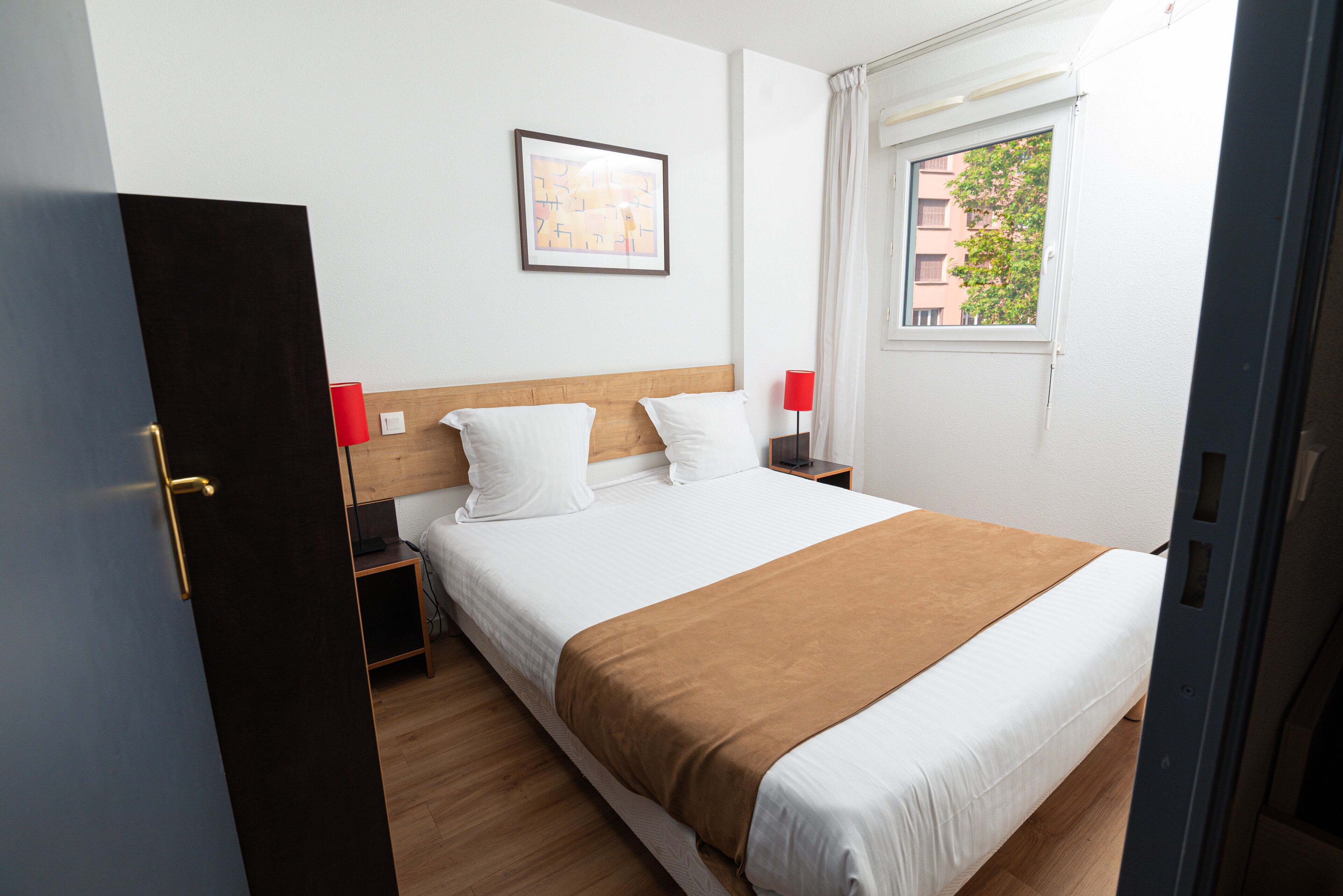 Una cama bien hecha con ropa de cama blanca y marrón, un cabecero de madera y una mesita de noche con una lámpara roja.
