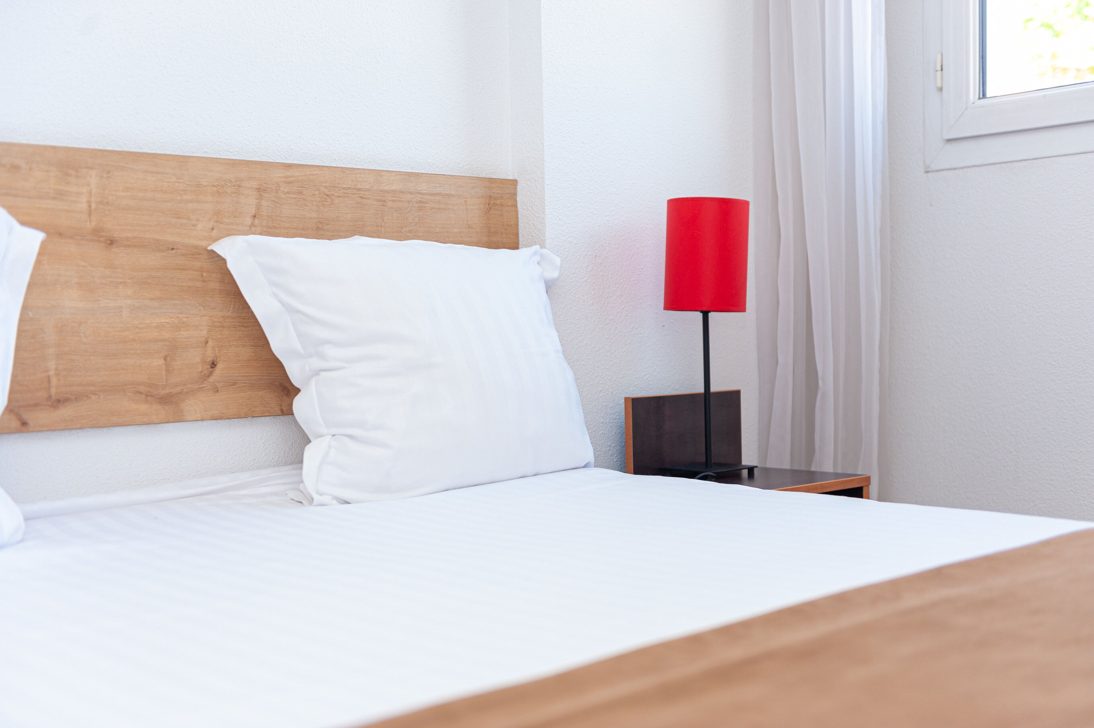 A neatly made bed with white linens and a wooden headboard, a red lamp on a bedside table, and a window with white curtains.