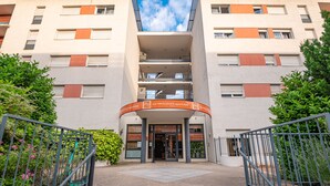 A multi-story hotel with a central entrance, outdoor seating area, and greenery. - Apparthôtel Privilodges Lyon (Lyon)