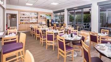 A restaurant with wooden chairs and tables, a kitchen area, and large windows.