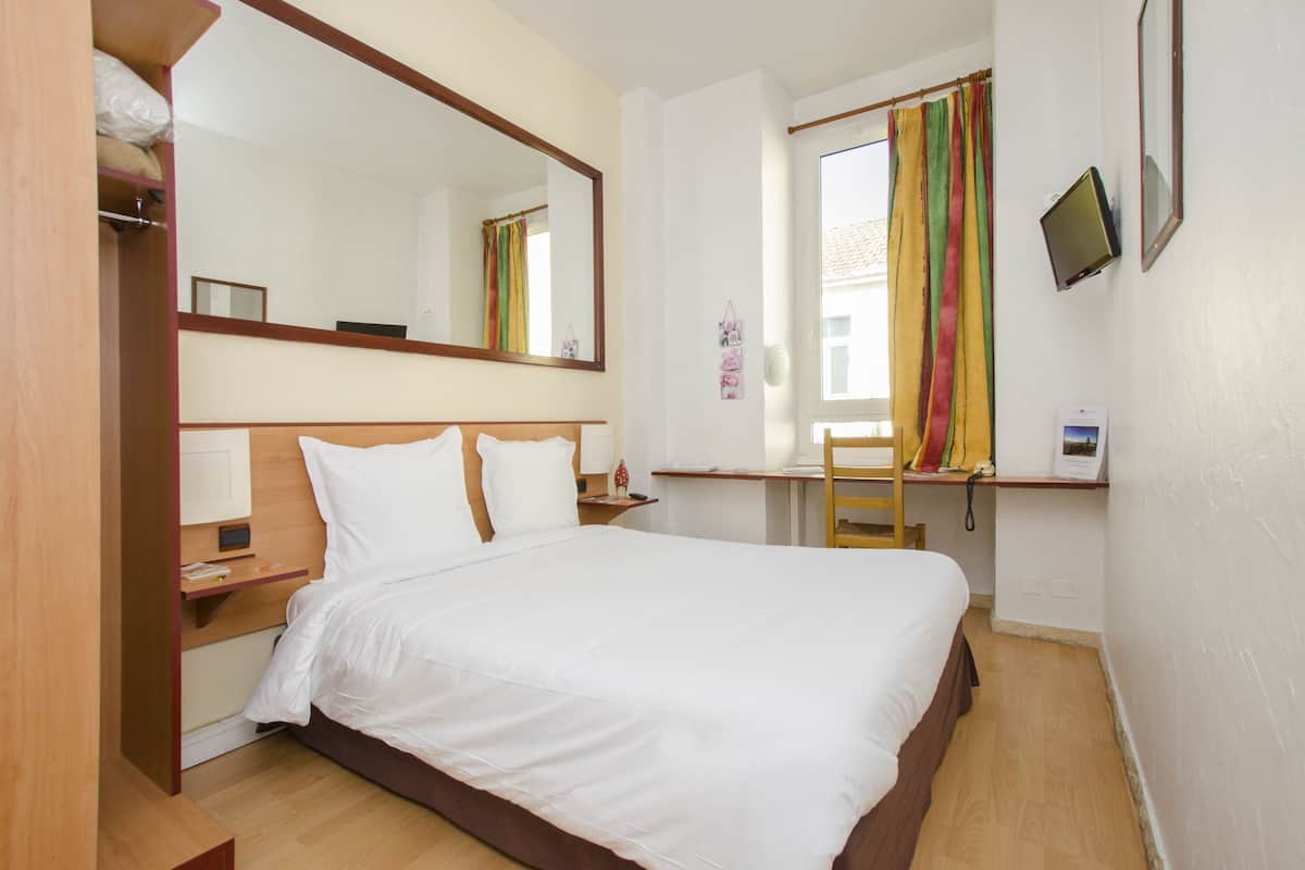 Une chambre d’hôtel avec un lit, un bureau, une chaise, un miroir et une fenêtre avec des rideaux.