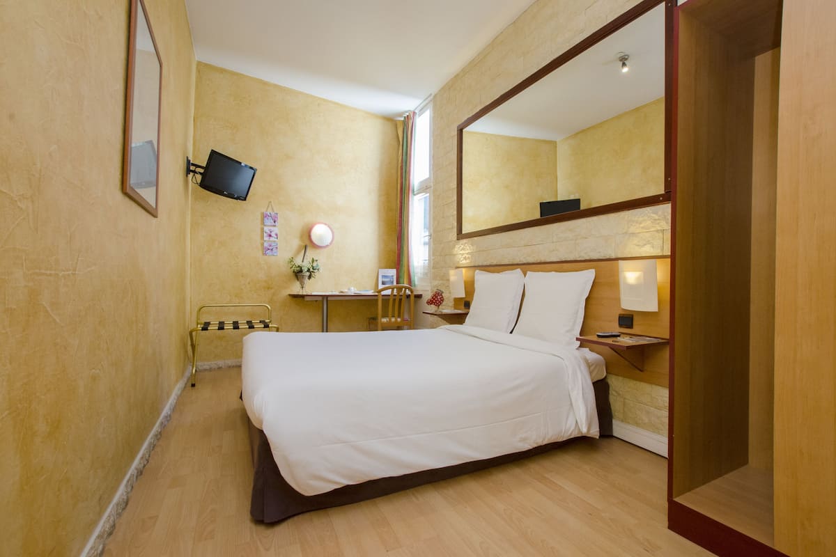 Une chambre d’hôtel comprenant un lit, une table de chevet, une lampe, un miroir, une télévision et un bureau avec une chaise.