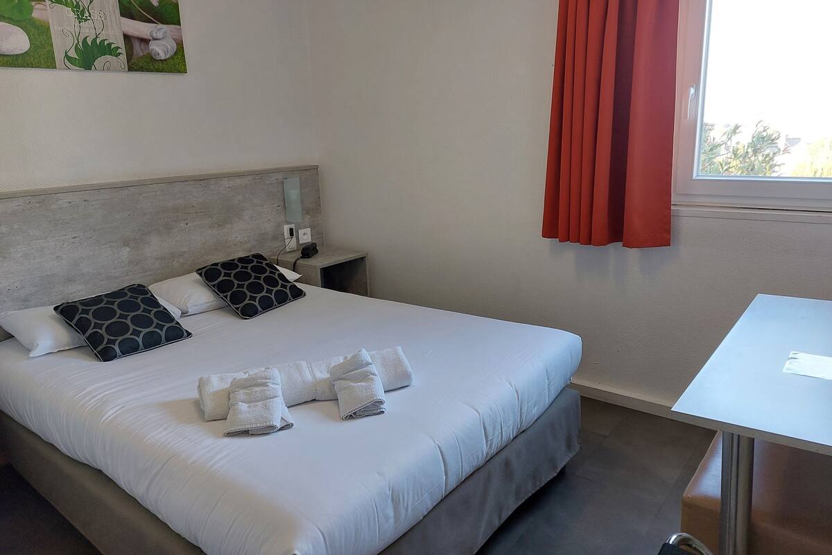 Un lit bien fait, avec du linge de lit blanc et deux coussins à motifs, une table de chevet avec un téléphone et une lampe, et une fenêtre avec des rideaux rouges.