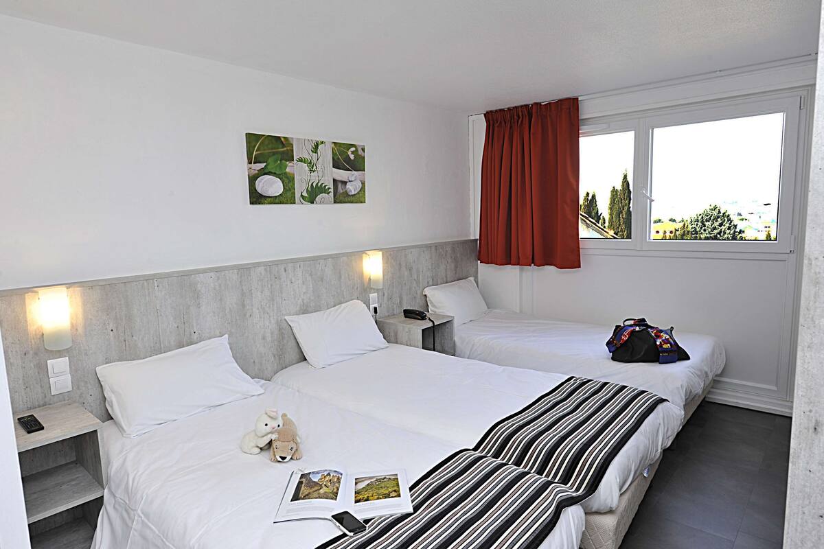 Une chambre d’hôtel avec deux lits, un rideau rouge, une vue sur la ville et une petite table sur laquelle se trouvent un ours en peluche et un livre.
