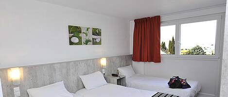 Een hotelkamer met twee bedden, een rood gordijn, uitzicht op de stad en een klein tafeltje met een teddybeer en een boek.