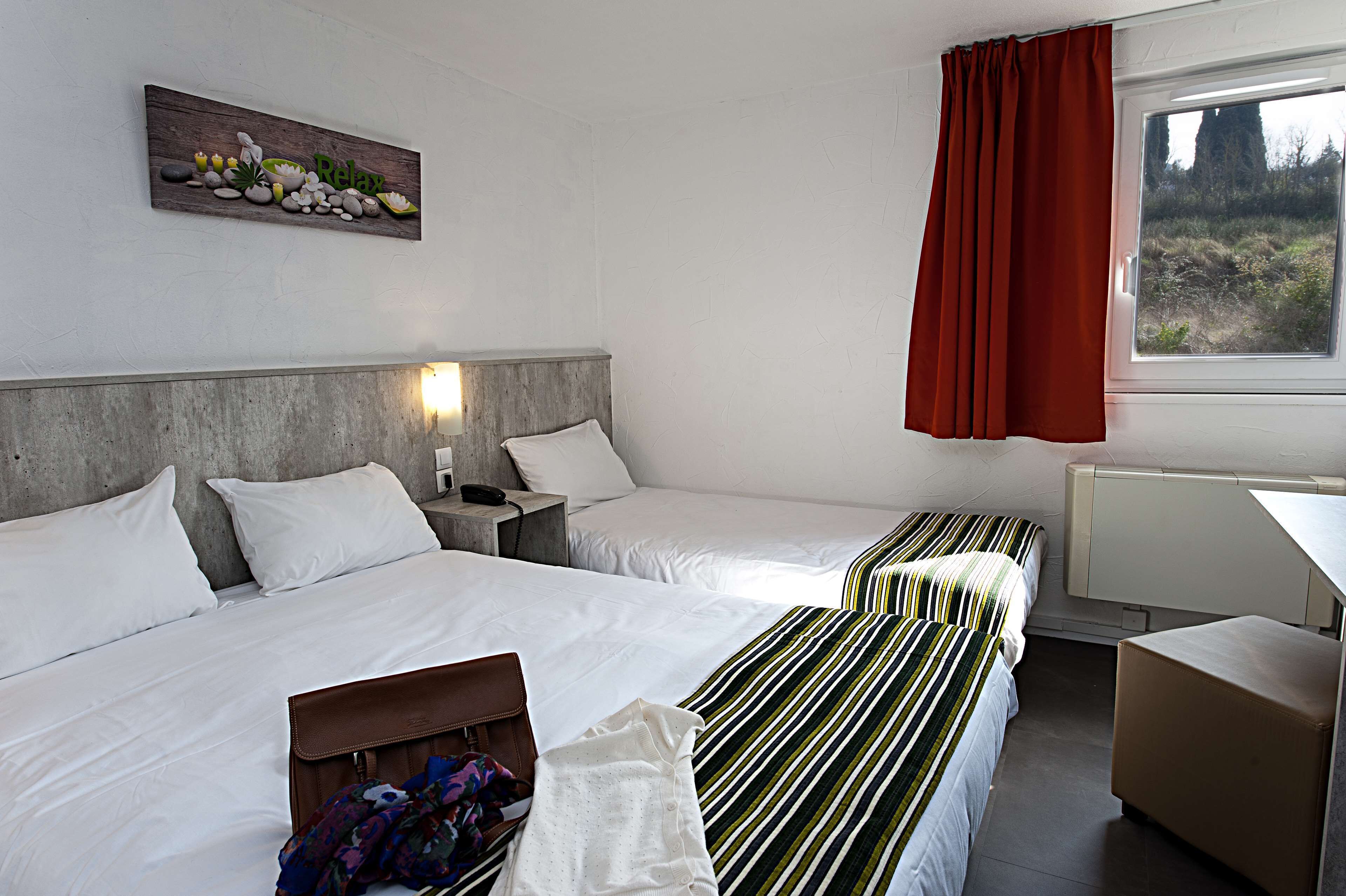 Quarto de hotel com duas camas, cabeceira de madeira, um quadro na parede, janela com cortinas vermelhas e uma luminária de cabeceira.