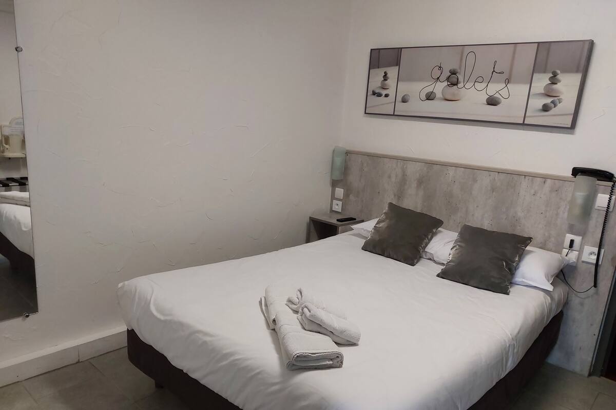 Un lit bien fait, avec du linge de lit blanc et deux oreillers, une table de chevet avec un téléphone, et une œuvre d’art accrochée au mur au-dessus du lit.
