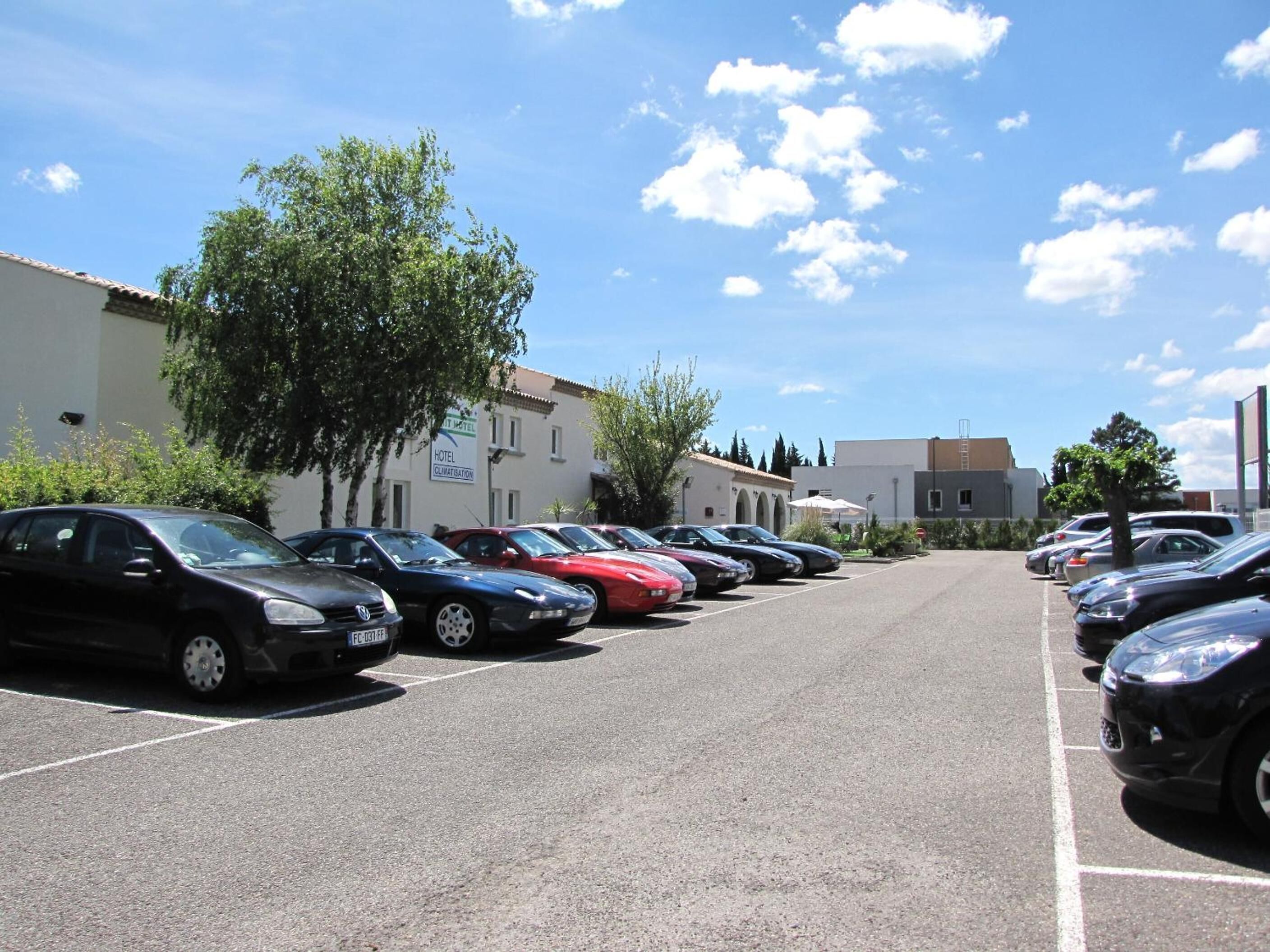 Un parking où plusieurs voitures sont garées en rangées, un bâtiment sur lequel on peut lire l’inscription « CLIMATISATION D’HÔTEL », et un ciel bleu clair parsemé de quelques nuages.