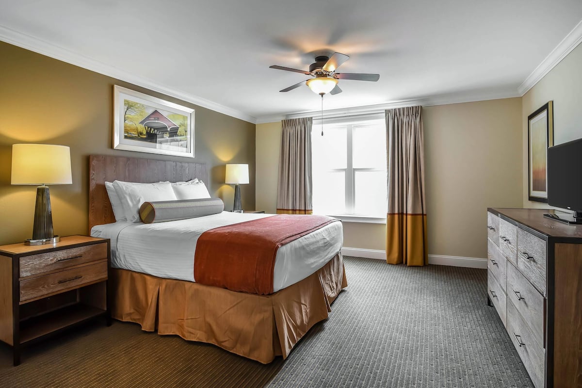 A hotel room with a bed, bedside lamps, a ceiling fan, a dresser, and a window with curtains.