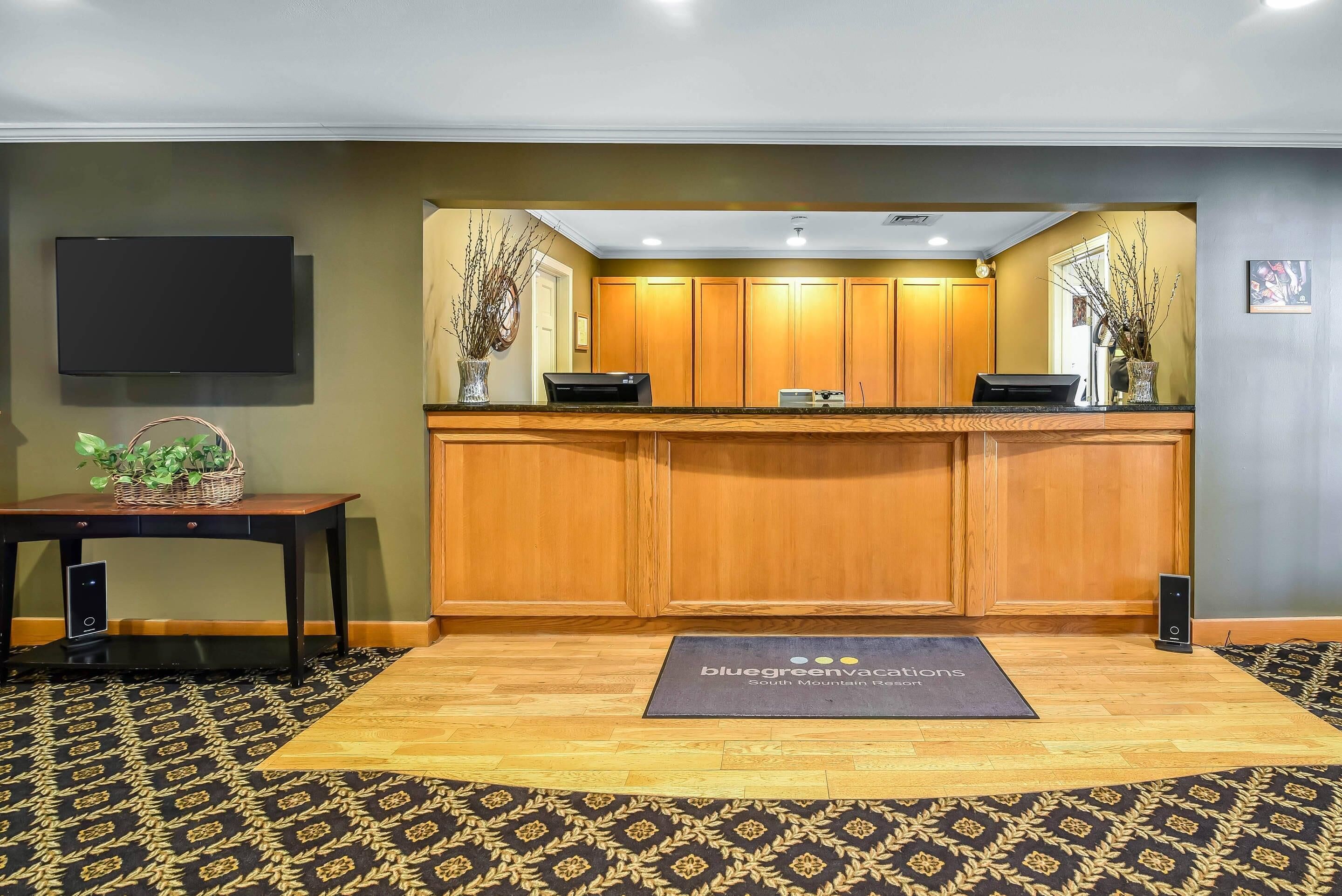 A reception area with a wooden counter, a flat-screen TV mounted on the wall, a reception desk, and a patterned carpet.