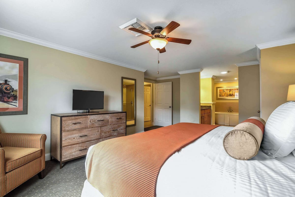 A hotel room with a bed, a television on a wooden dresser, a ceiling fan, and a door leading to a bathroom.