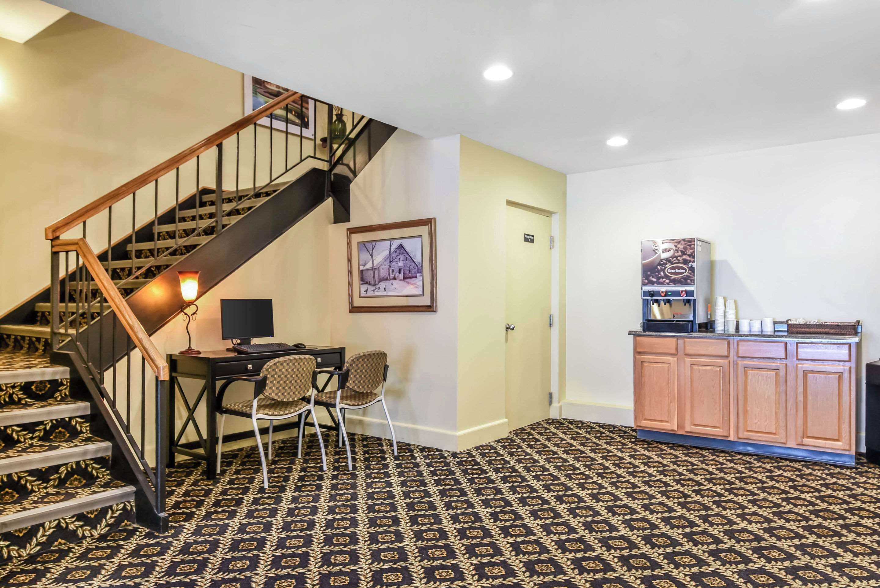A hotel lobby with a staircase, a desk with a computer, a chair, a framed picture on the wall, and a coffee station.