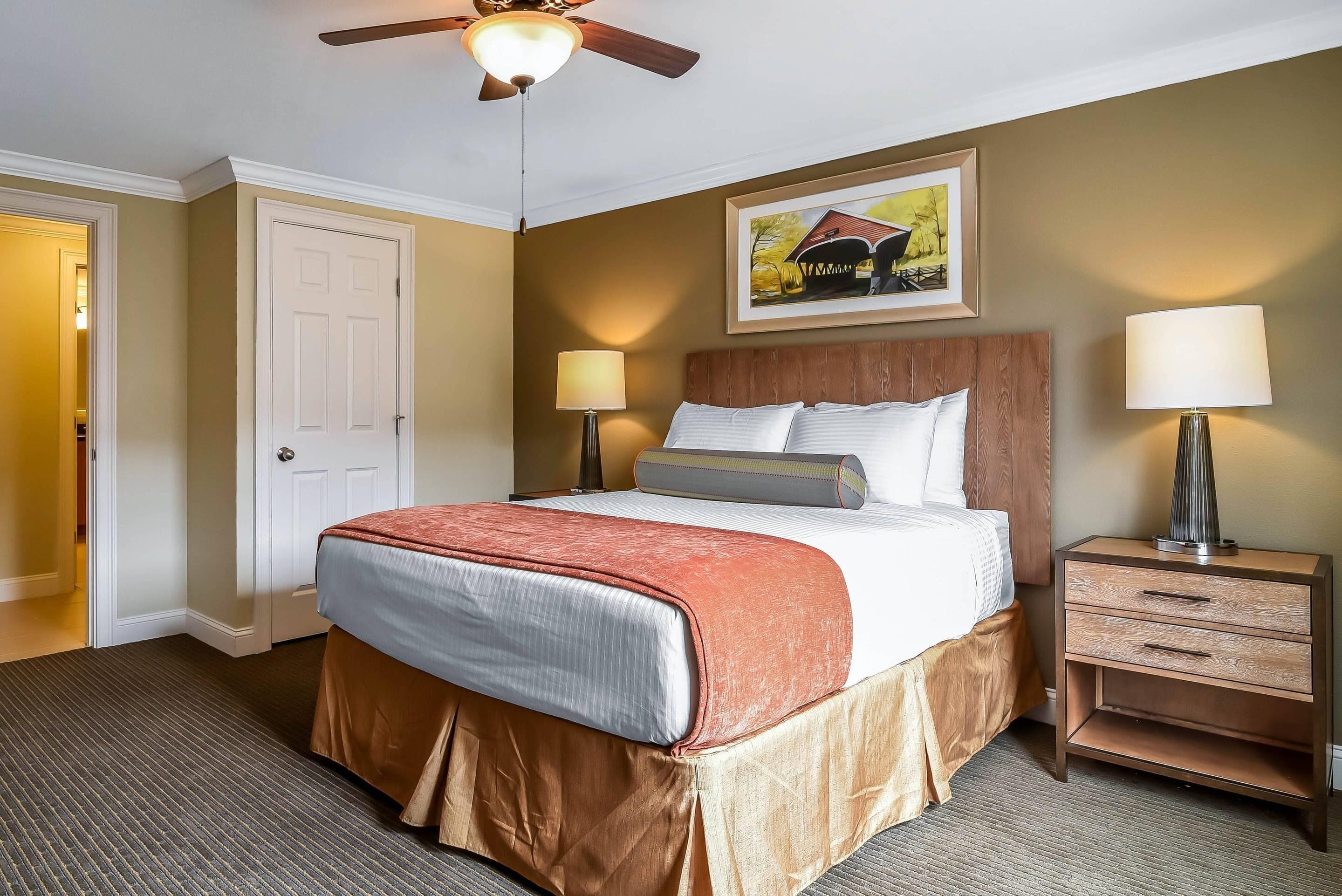 A hotel room with a bed, two bedside lamps, a nightstand with drawers, a ceiling fan, and a framed picture on the wall.