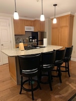 Una cocina con armarios de madera, microondas, refrigerador, encimera de granito y cuatro taburetes negros.