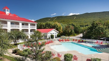 Un hotel con piscina, edificio de techo rojo y montañas al fondo.