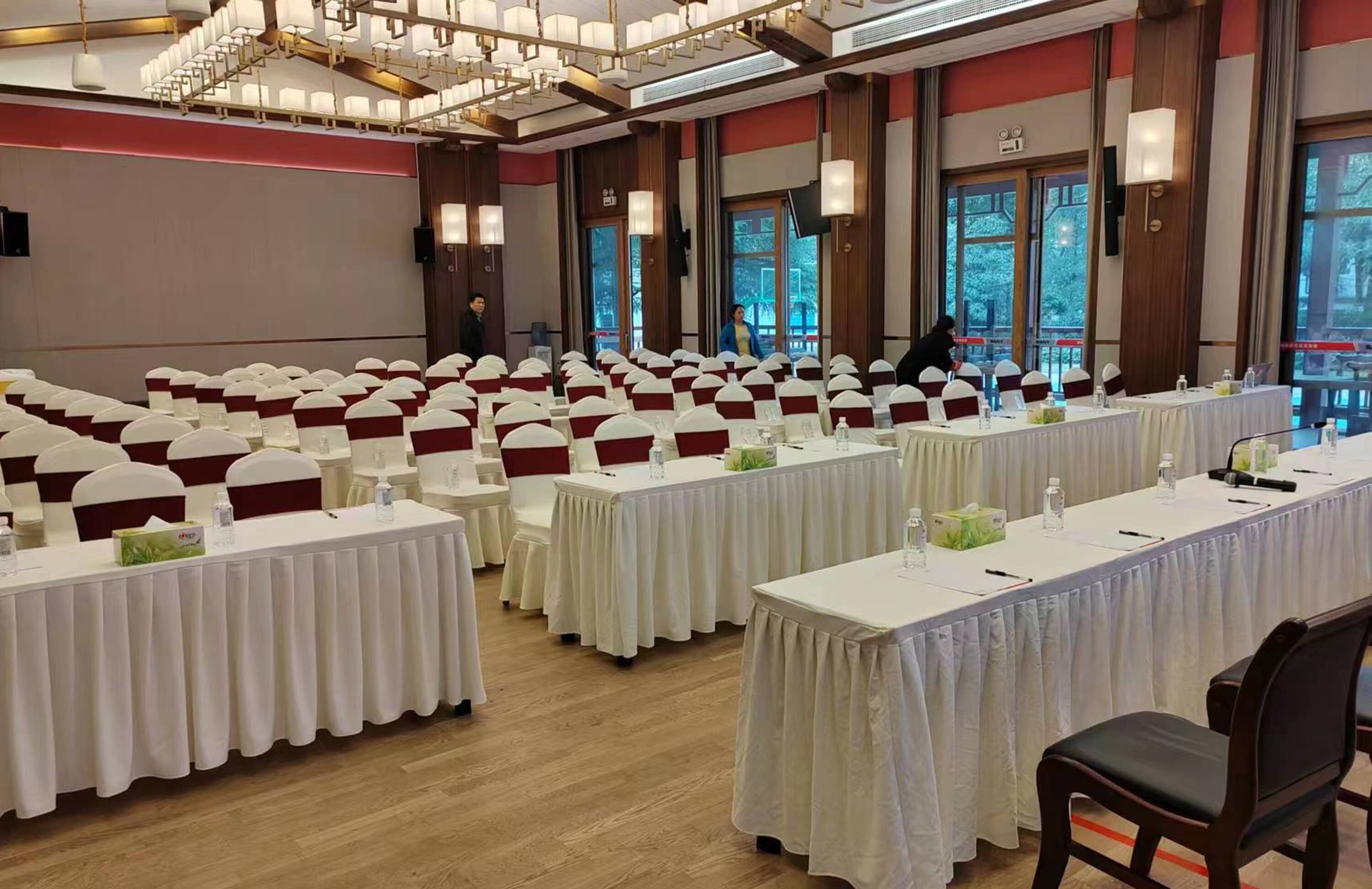 Un salón de conferencias con filas de mesas, cada una con mantel blanco y cintas rojas, y un ventanal con vistas a árboles.