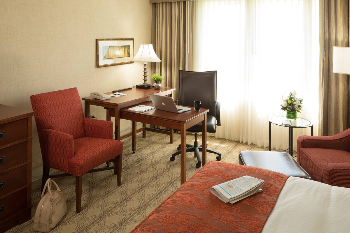 A hotel room with a bed, two red armchairs, a desk with a laptop, a chair, a TV, a dresser, and a small table with a vase of flowers.