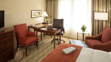 A hotel room with a bed, two red armchairs, a desk with a laptop, a chair, a TV, a dresser, and a small table with a vase of flowers.