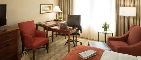 A hotel room with a bed, two red armchairs, a desk with a laptop, a chair, a TV, a dresser, and a small table with a vase of flowers.