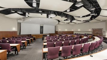 A conference room with rows of purple chairs, a large white screen, and a curved table in the center.