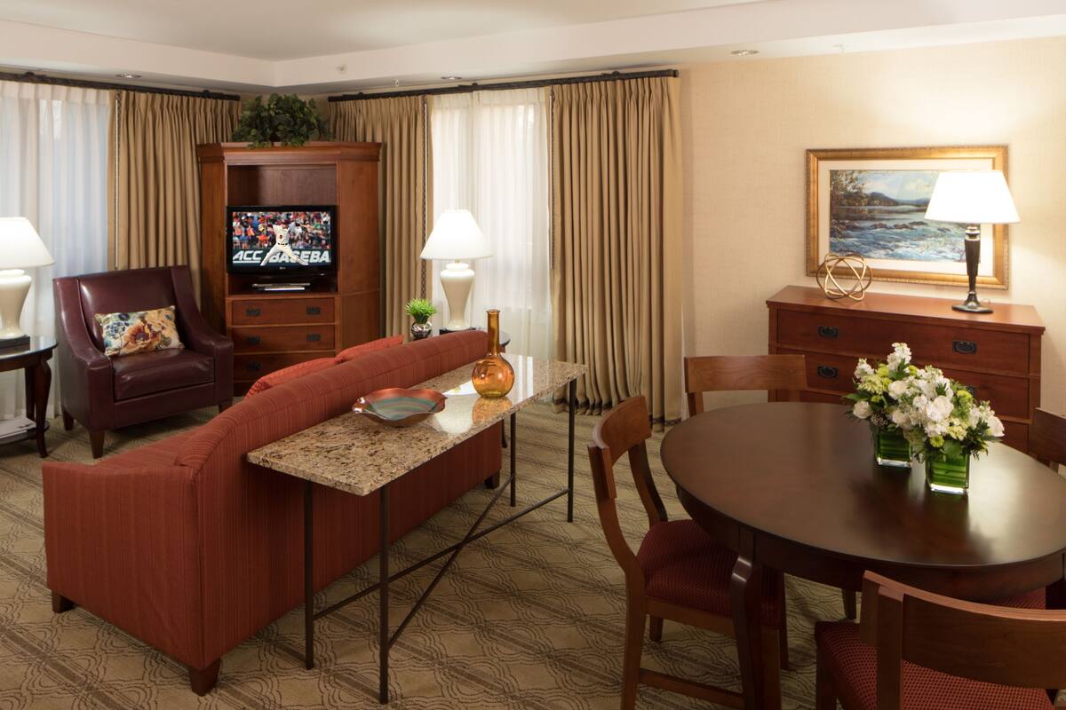 A hotel room with a living area, a dining table with chairs, a television, a sofa, a coffee table, a lamp, a painting, and a plant.
