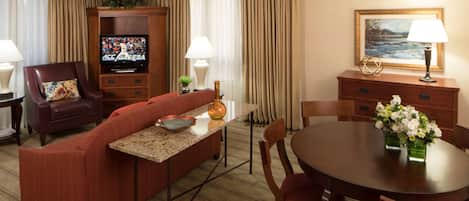 Ein Hotelzimmer mit Wohnbereich, einem Esstisch mit Stühlen, einem Fernseher, einer Couch, einem Couchtisch, einer Lampe, einem Gemälde und einer Pflanze.