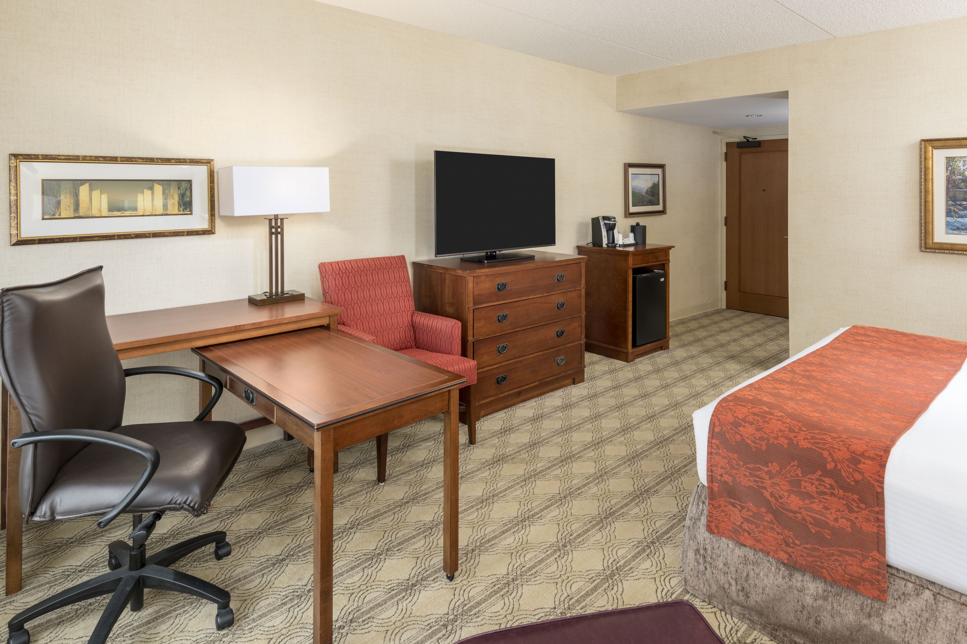 Habitación de hotel con cama, escritorio y silla, televisión, cómoda y puerta que da a otra habitación.