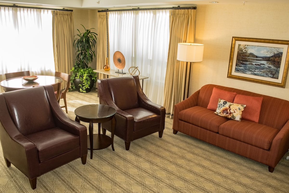 A hotel room with a brown leather sofa, two leather armchairs, a small round table, a potted plant, a lamp, and a framed picture on the wall.