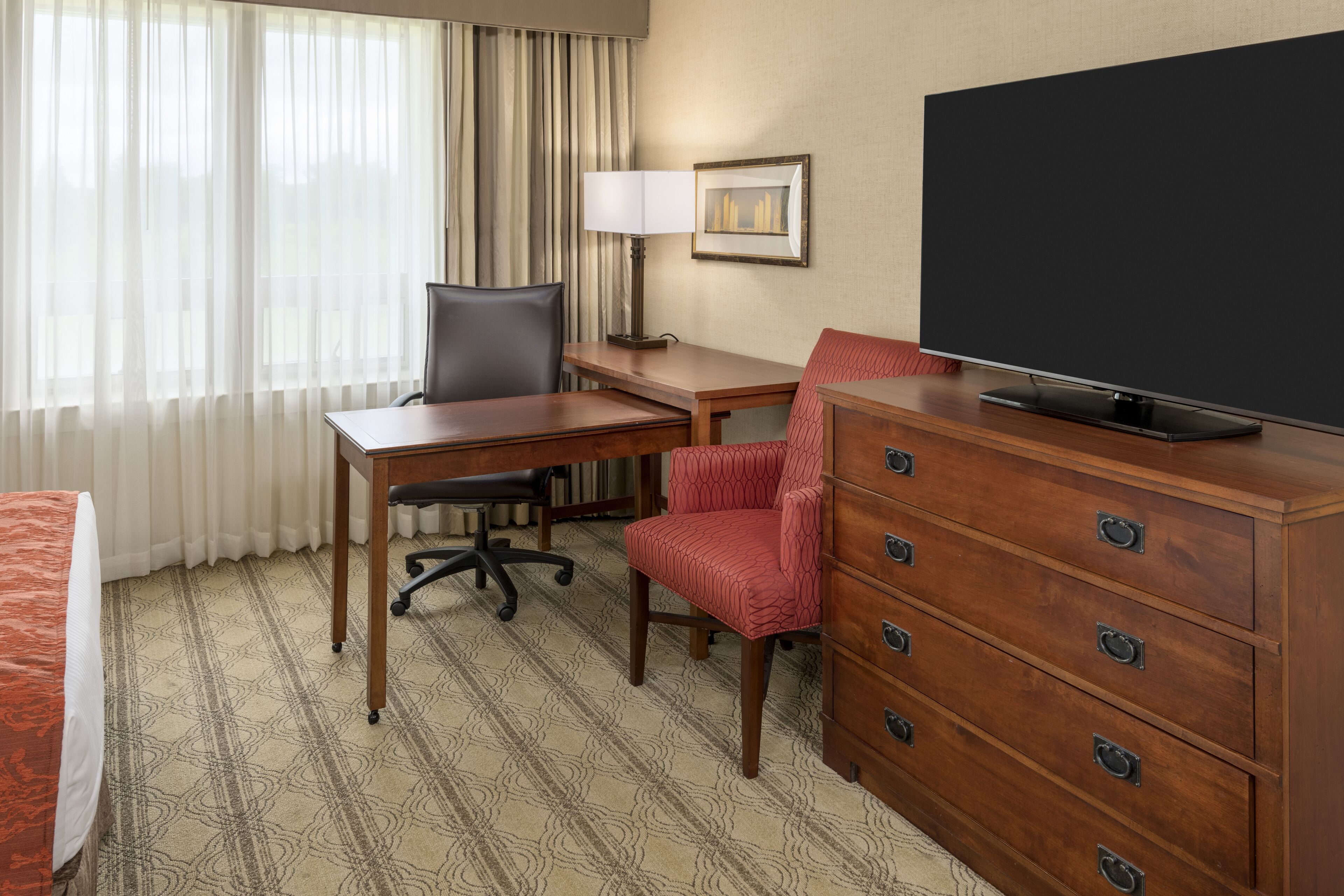 A hotel room with a bed, a desk, a chair, a dresser, a television, and a window with curtains.