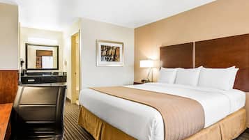 A hotel room with a bed, a desk, a chair, a mirror, a painting, and a nightstand with a lamp.