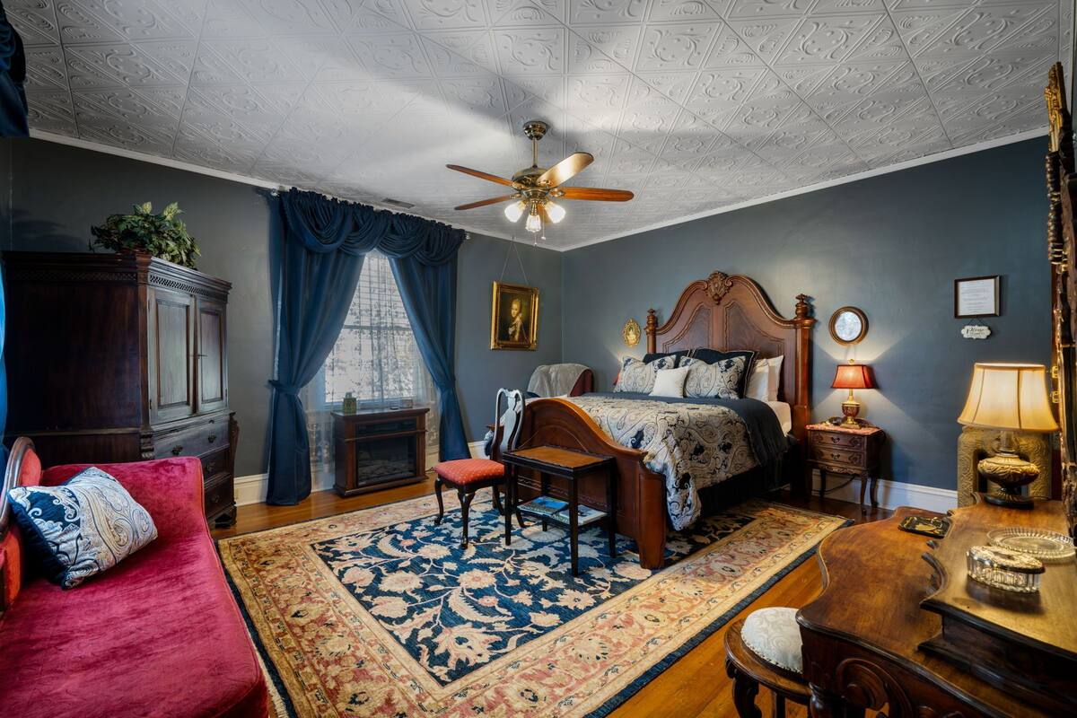 A bedroom with a wooden bed, a red sofa, a wooden desk, a ceiling fan, and a large window with blue curtains.