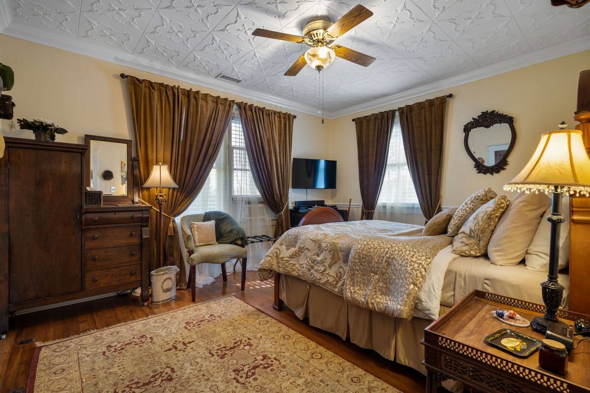 A bedroom with a bed, a desk, a chair, a television, and a ceiling fan.