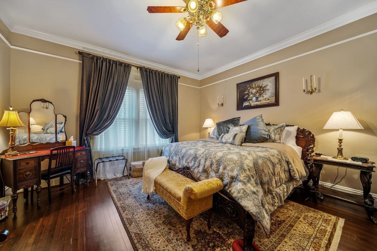 A bedroom with a large bed, a desk, a chair, a bench, a ceiling fan, and a window with curtains.