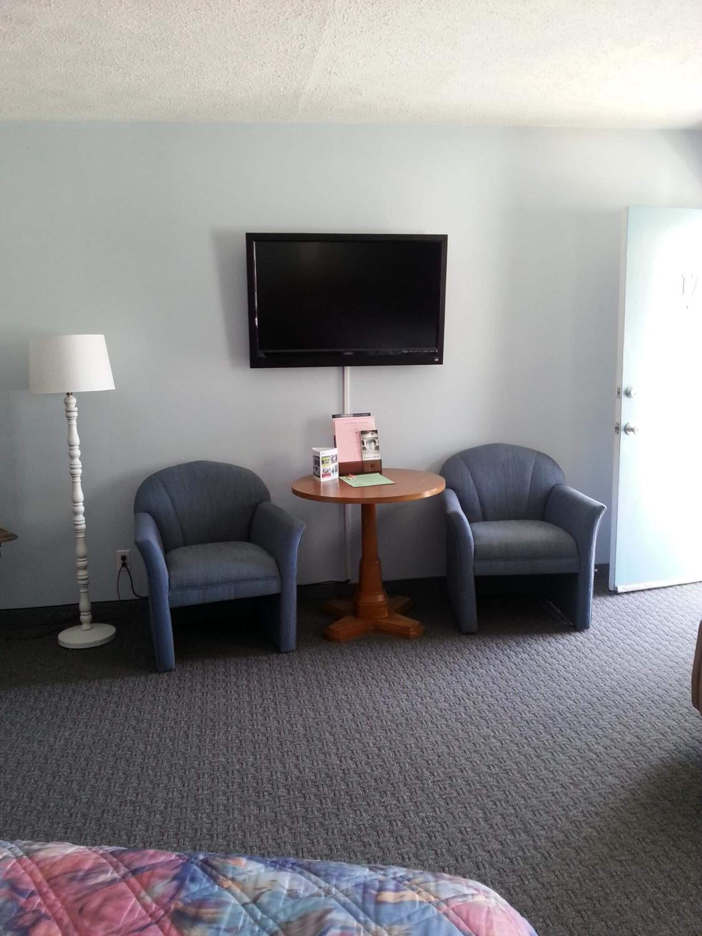 Une chambre d’hôtel avec deux fauteuils bleus, une table ronde en bois, un lampadaire blanc et un téléviseur à écran plat fixé au mur.