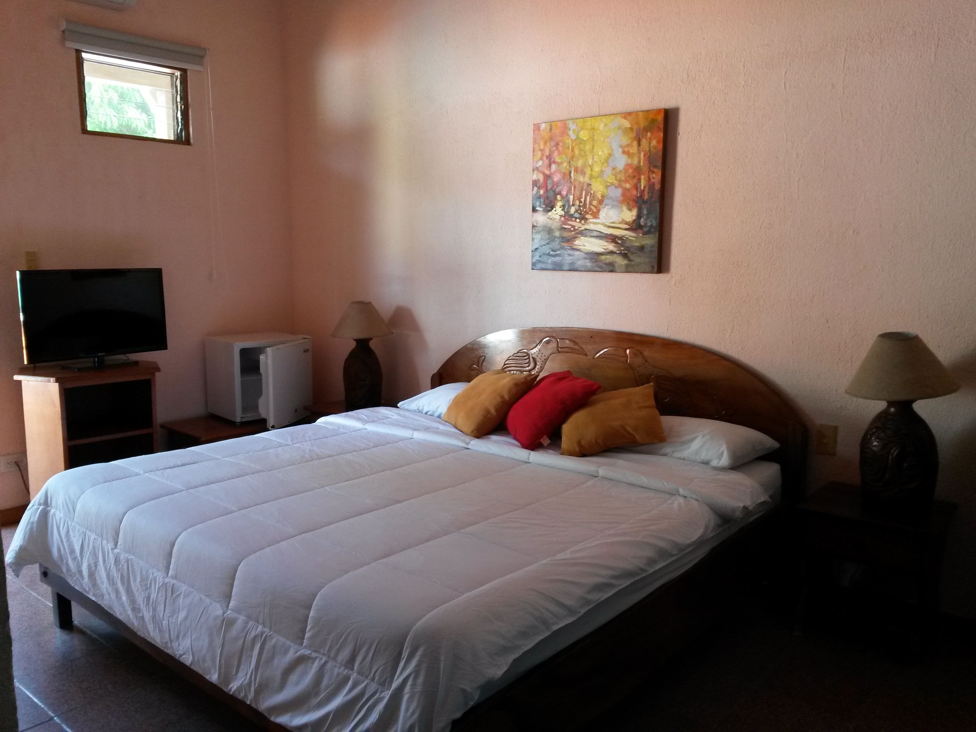 A neatly made bed with a white duvet, a painting on the wall, a television on a stand, and two lamps on either side of the bed.