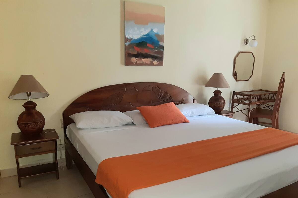 Een net opgemaakt bed met een oranje deken, twee bedlampen, een houten nachtkastje, een houten stoel en een schilderij aan de muur.