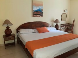 Una cama bien hecha con una manta naranja, dos lámparas de noche, una mesita de madera, una silla de madera y un cuadro en la pared.