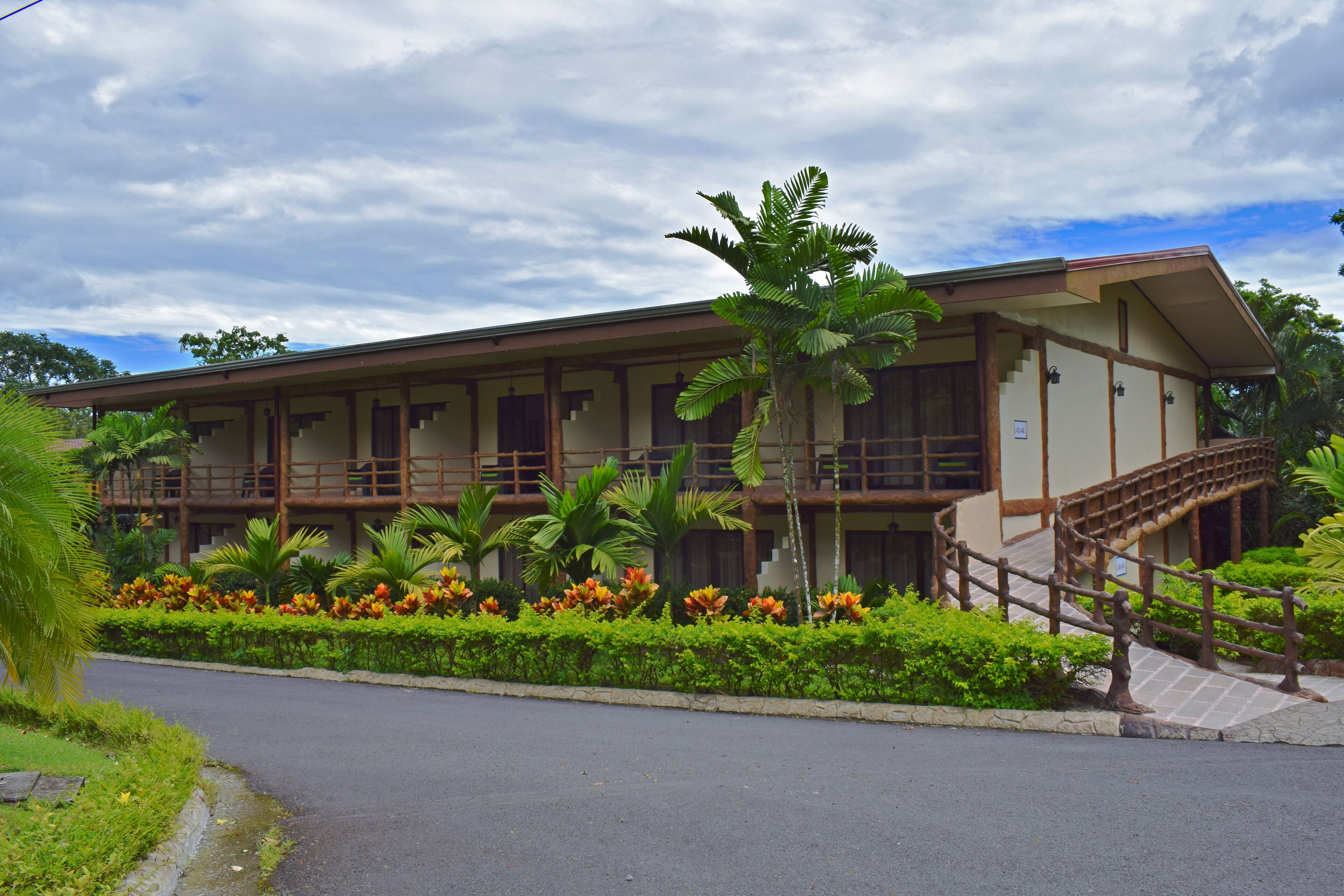 A two-story building with a balcony, a staircase, and a garden with tropical plants.