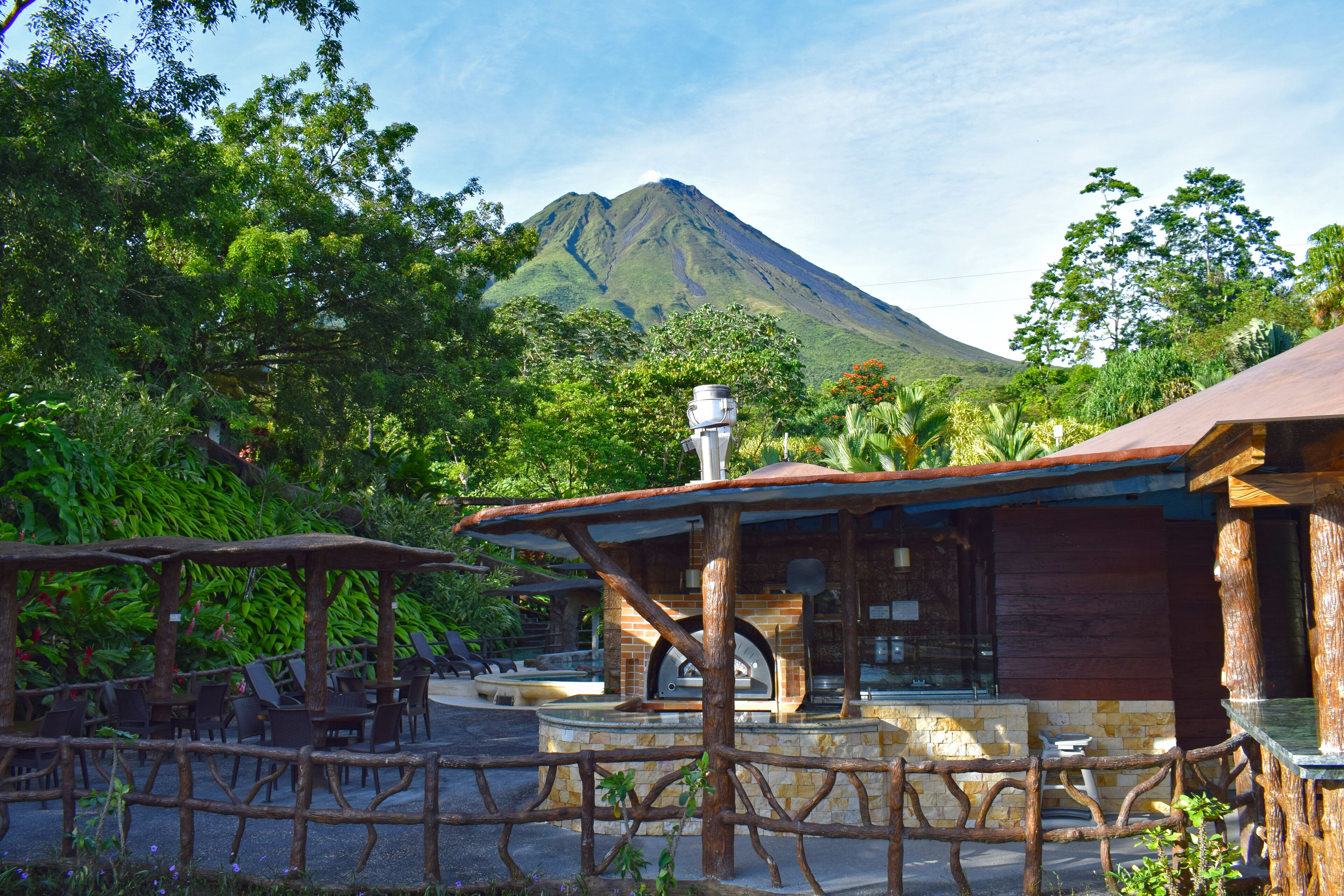 A rustic outdoor seating area with wooden tables and chairs, a stone fireplace, and a view of a mountain.