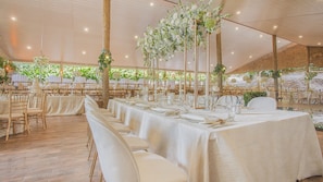 A tented event space with tables set for a formal gathering, featuring white tablecloths and floral centerpieces.