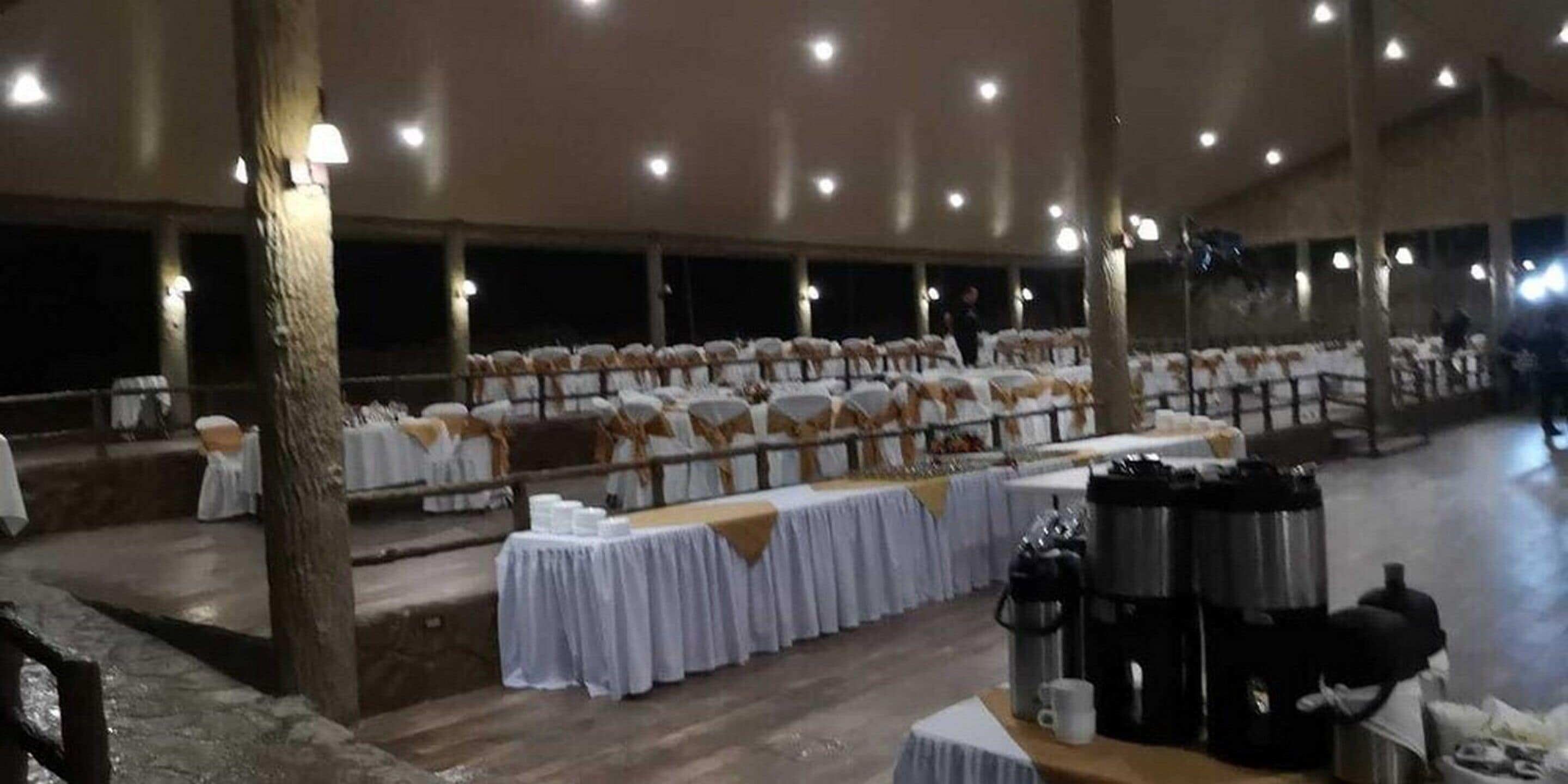 A banquet hall with rows of tables covered in white tablecloths, chairs with white and gold sashes, and a coffee station.