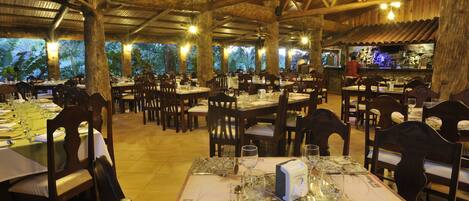Un comedor interior con mesas y sillas de madera, una zona de bar con mostrador de piedra y vistas a palmeras a través de amplios ventanales.