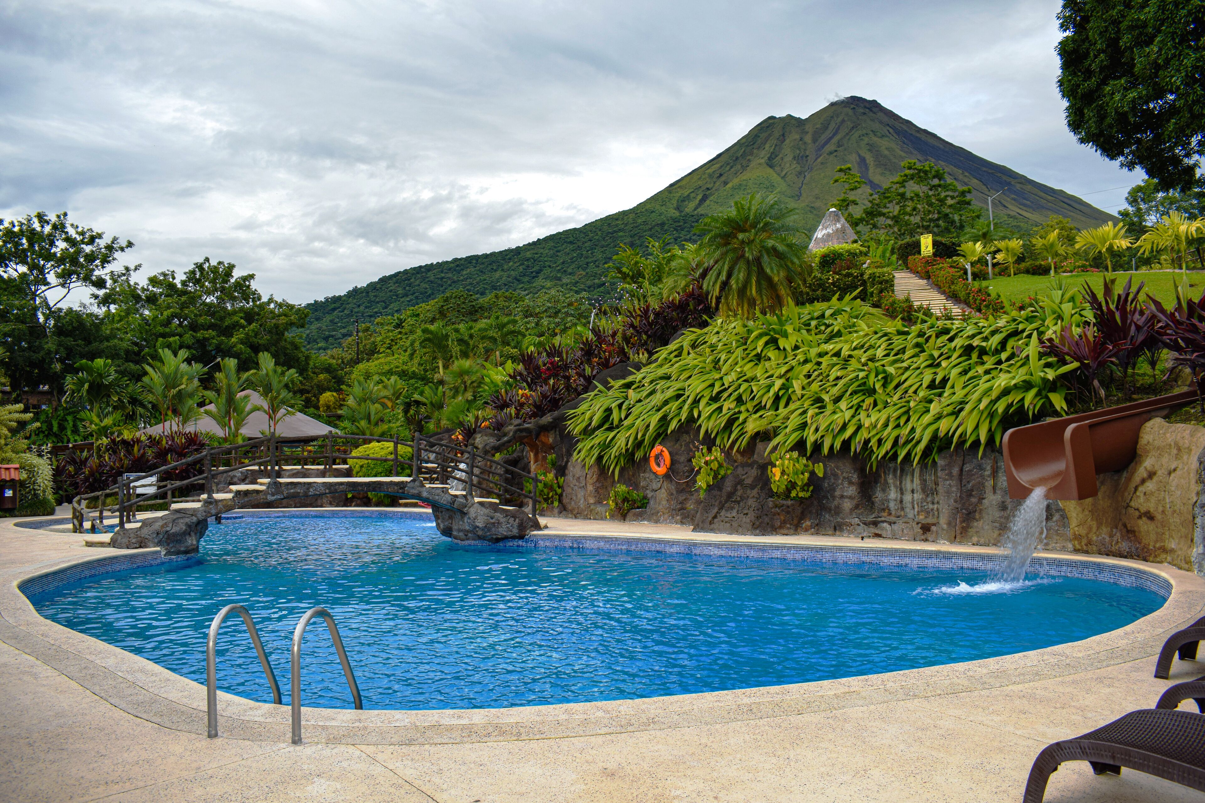 A swimming pool with a waterfall feature, surrounded by lush greenery and a mountain in the background.