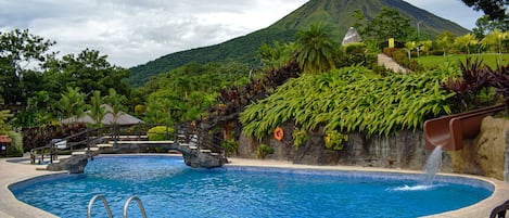 A swimming pool with a waterfall feature, surrounded by lush greenery and a mountain in the background.