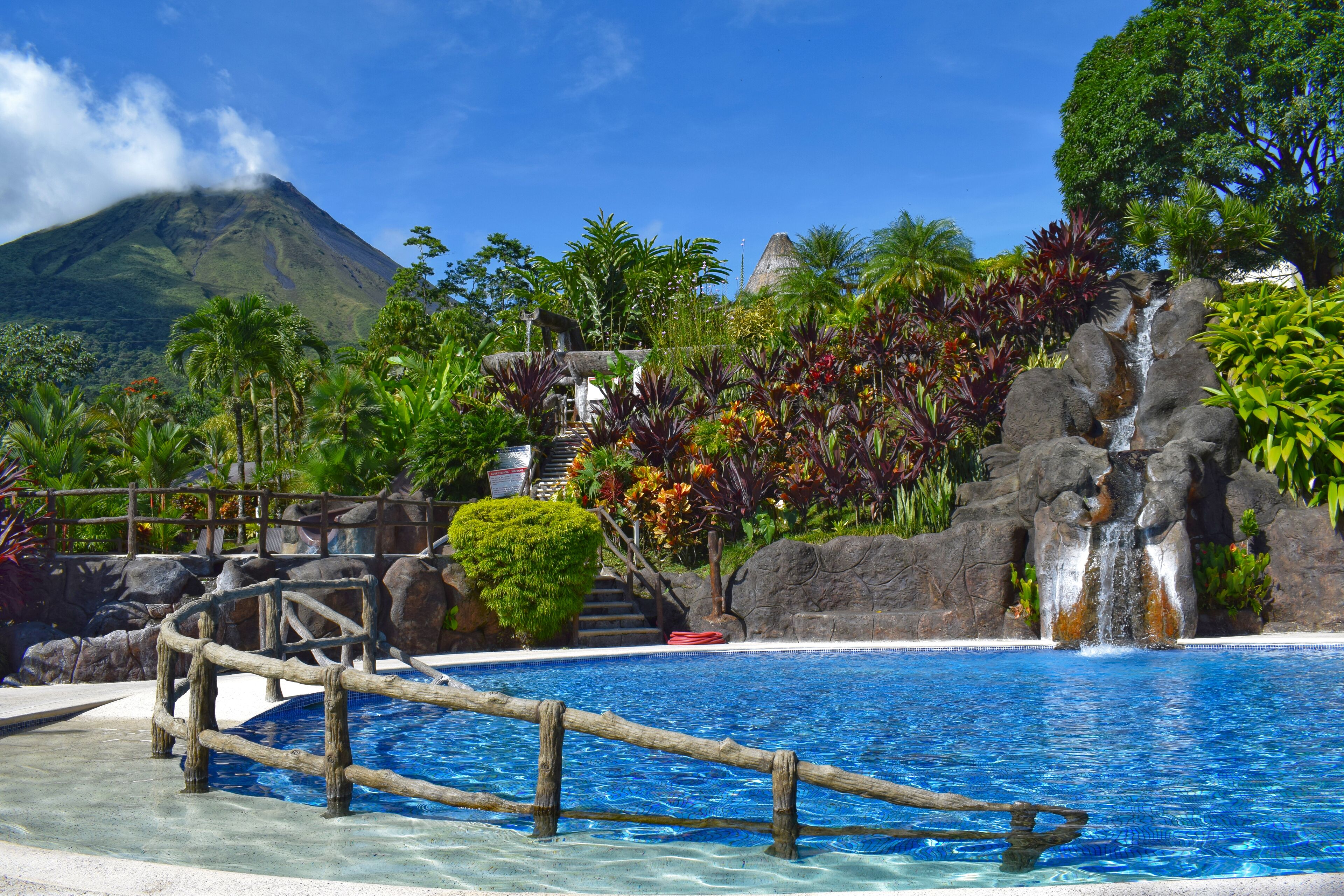 A pool area with a wooden bridge, a waterfall, and a mountain in the background.