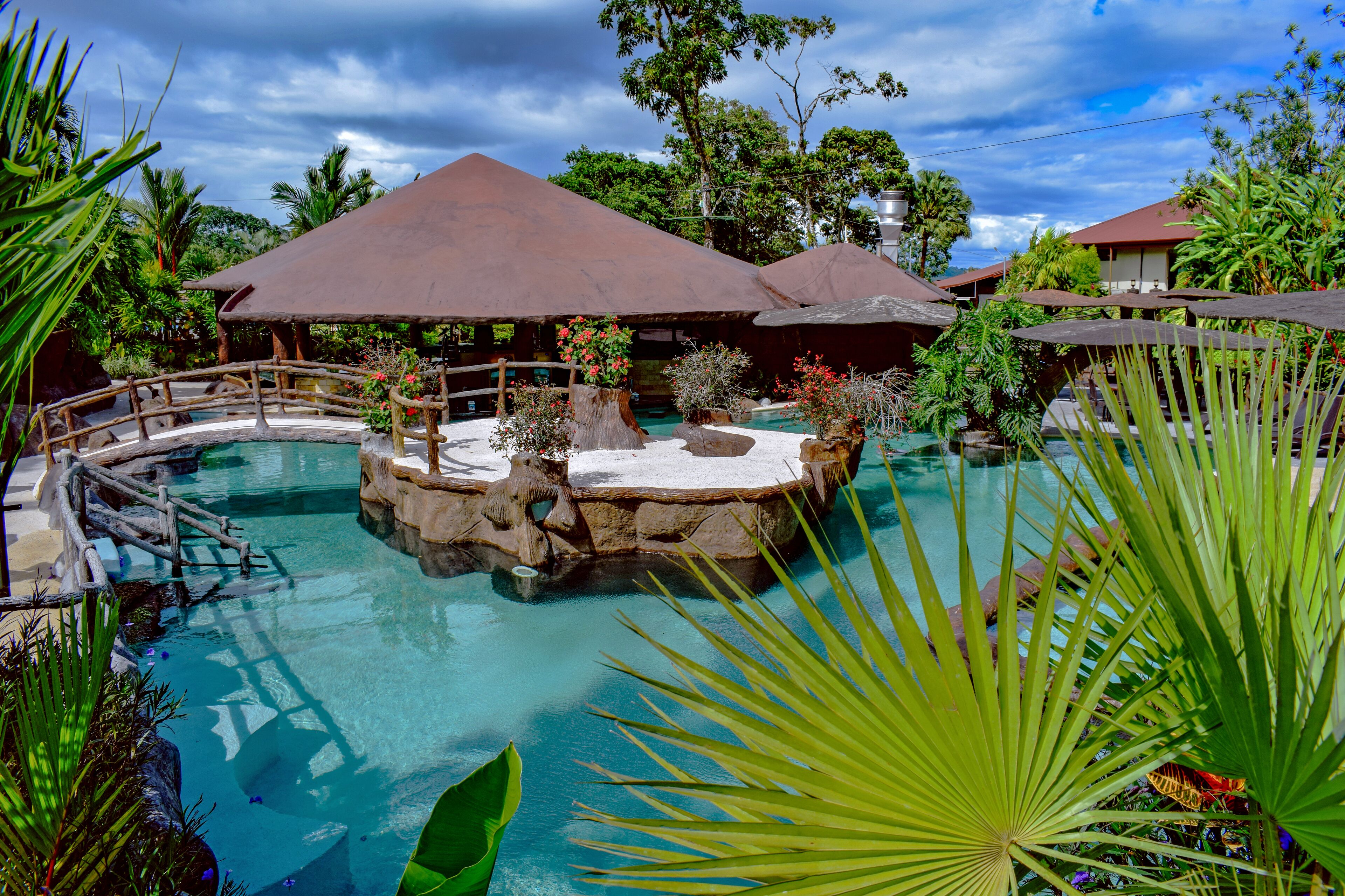 A tropical resort with a central pool, surrounded by lush greenery and covered seating areas.