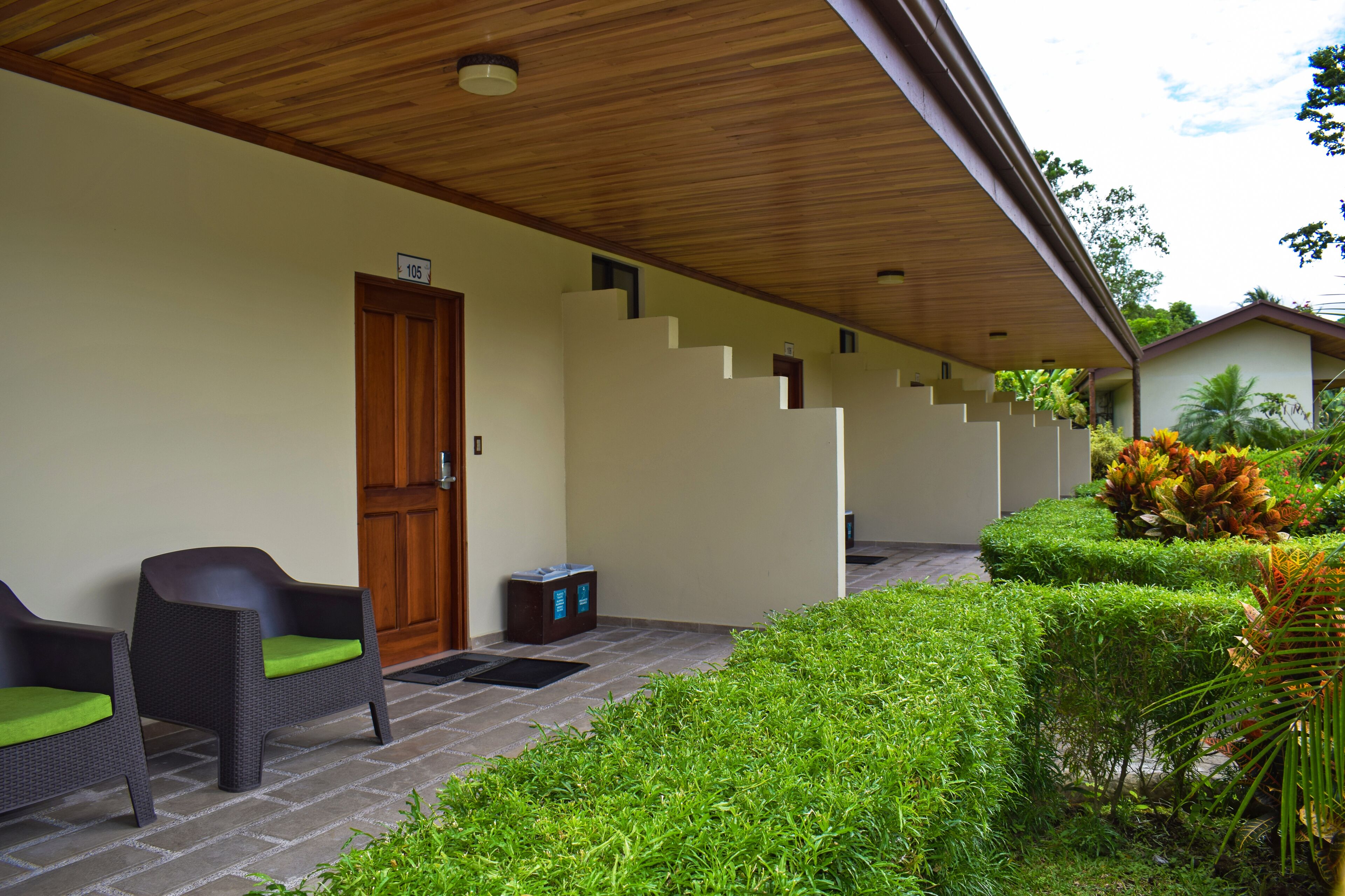 A covered outdoor area with a wooden ceiling, a door labeled '105', and two wicker chairs with green cushions.